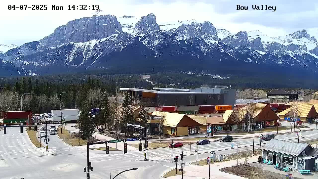 A mountain range capped with snow is visible in the background of a town scene with several buildings, vehicles, and roads; the weather is clear and bright.
