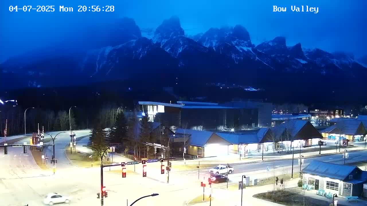 A nighttime view of a town at the base of snow-capped mountains under a dark blue twilight sky.