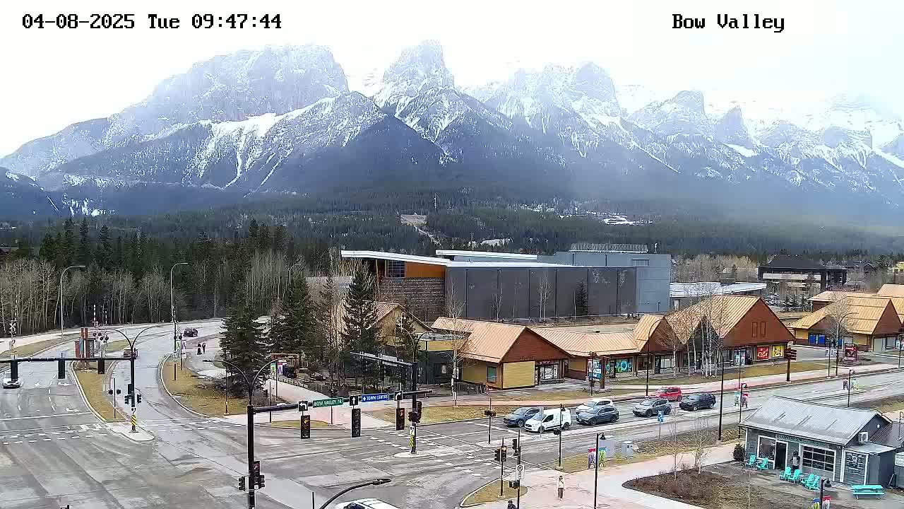A partly cloudy day reveals a mountain range with snow-capped peaks behind a town with roads, shops, and buildings.