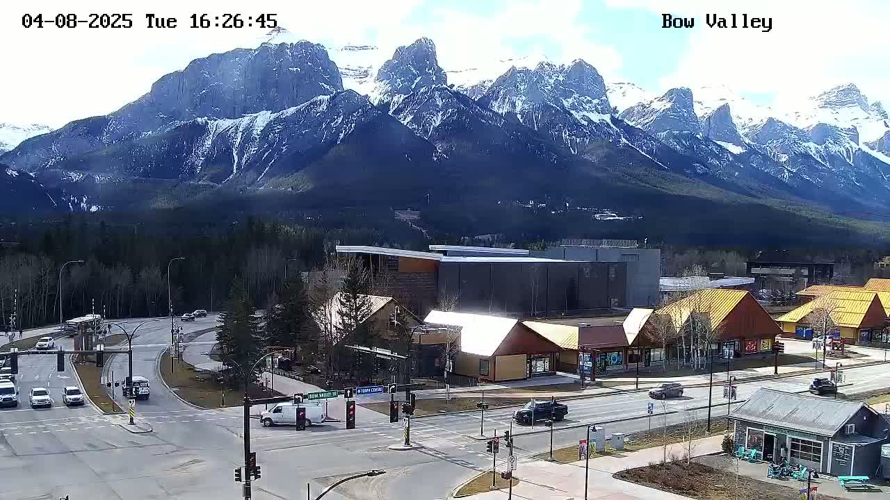 A partly sunny day shows a mountain range capped with snow behind a town with several buildings and vehicles at an intersection.