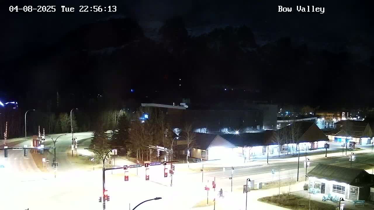 A nighttime, snow-covered roadway scene shows a crossroads with buildings and streetlights, with dark mountains in the background under a clear sky.