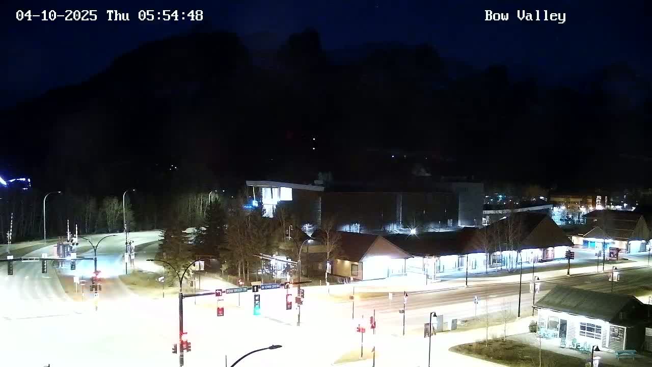 A nighttime view of a snow-covered intersection and buildings, with dark mountains in the background under a clear sky.