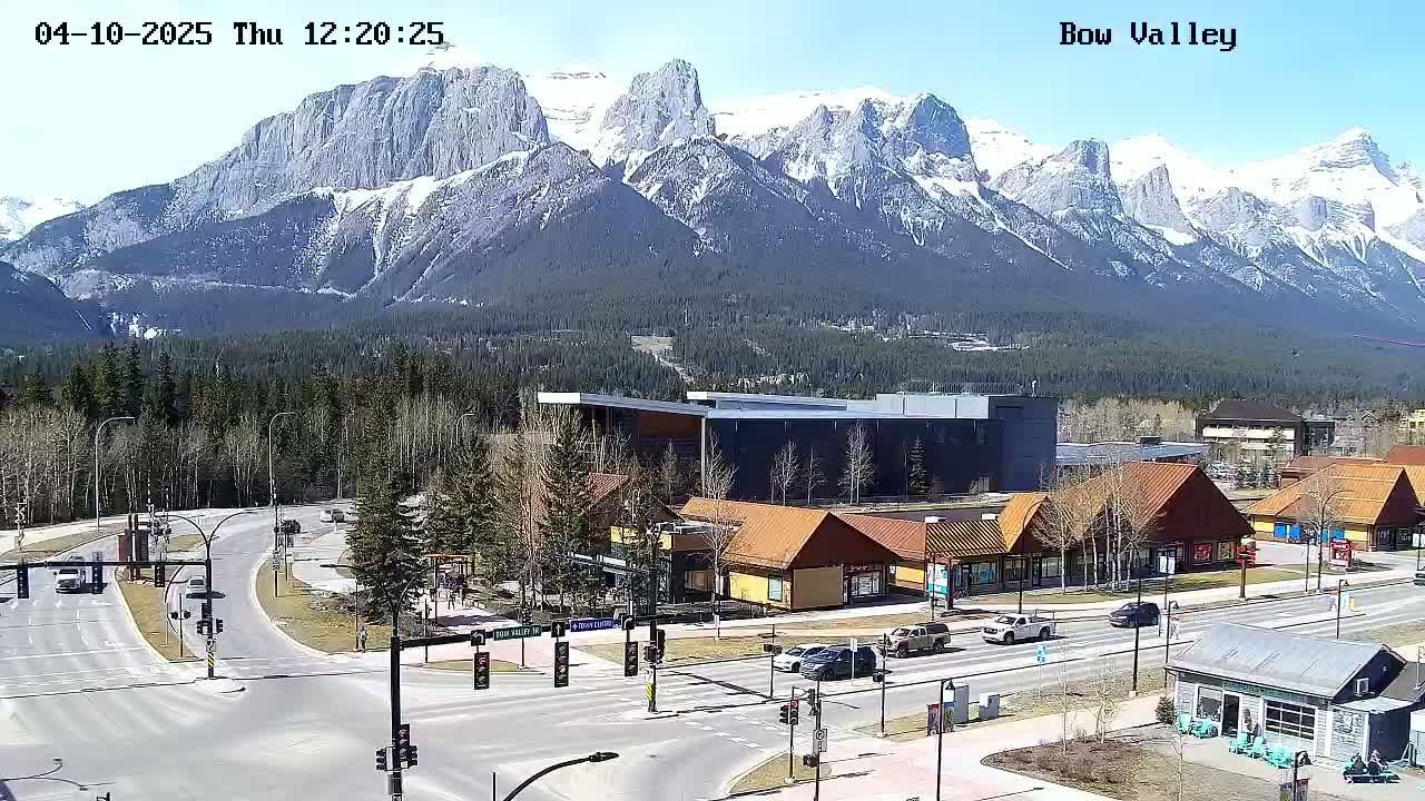 A sunny view of a mountain range with snow-capped peaks overlooking a town with buildings and roads, with several cars visible.