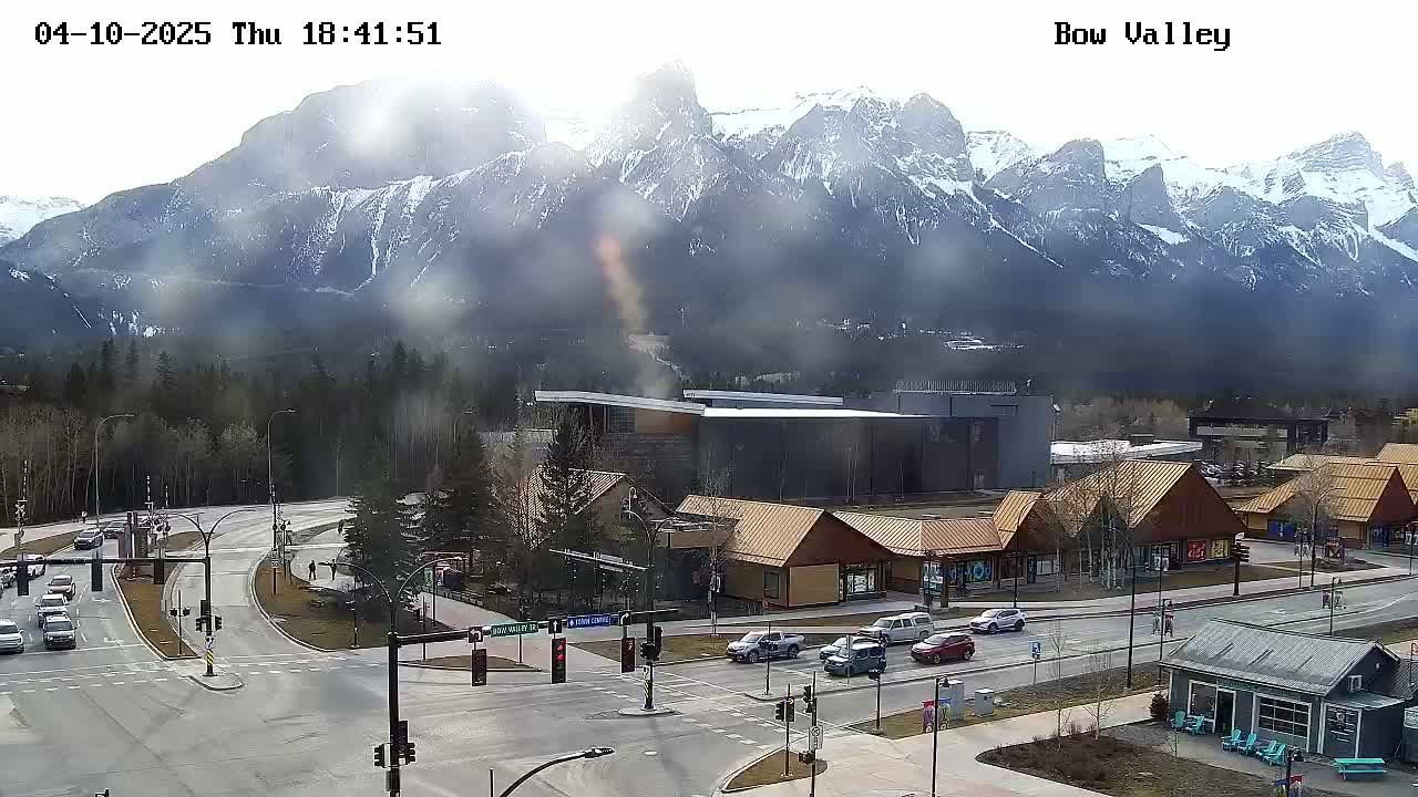 A hazy, sunny day reveals snow-capped mountains behind a mountain town intersection with several small buildings and cars.