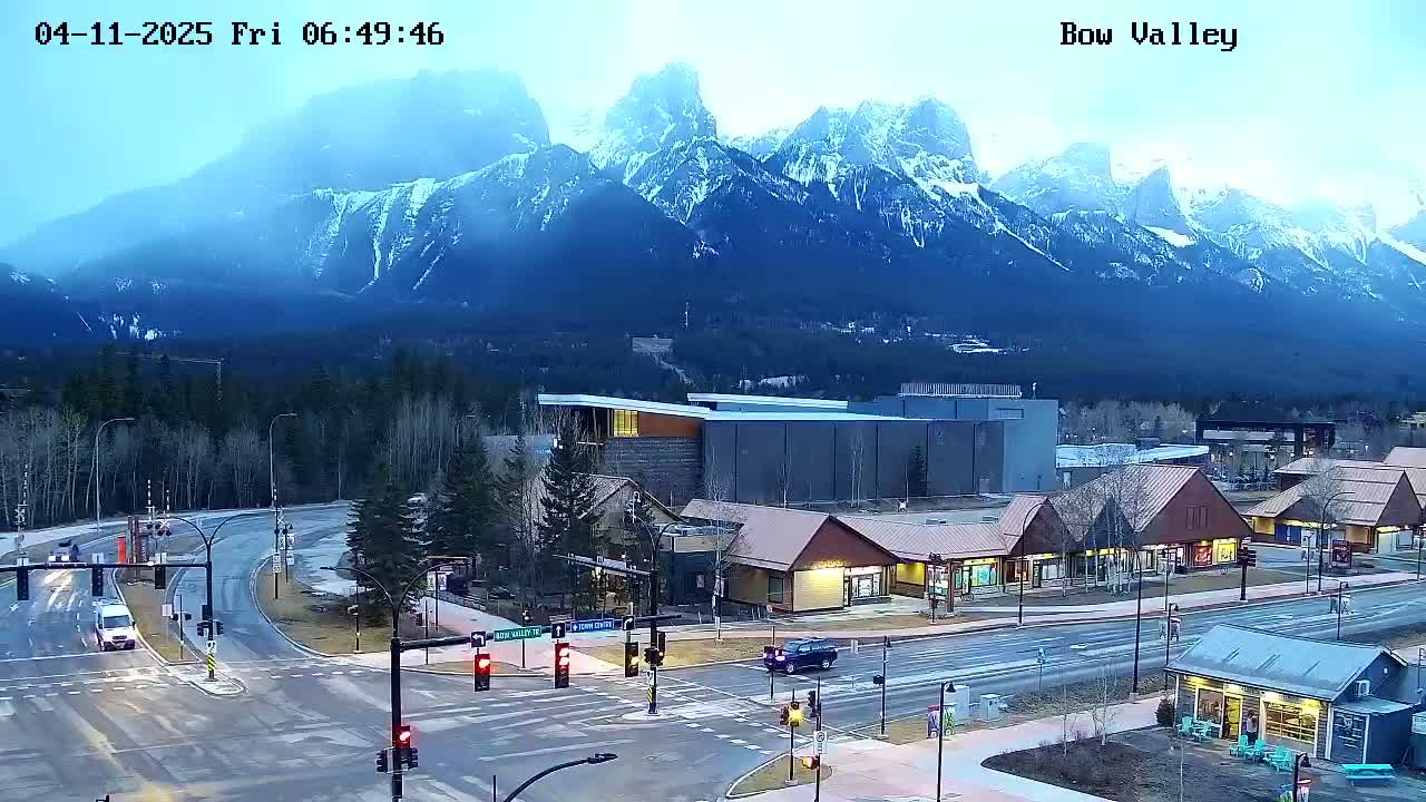 A mountain range capped with snow is visible in the background of a wet, overcast town with a few cars on the road.