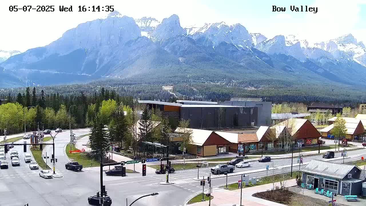 A mountain range with some snow on the peaks is visible in the background of a sunny outdoor scene showing a road intersection with several cars and buildings, including shops and a larger building.