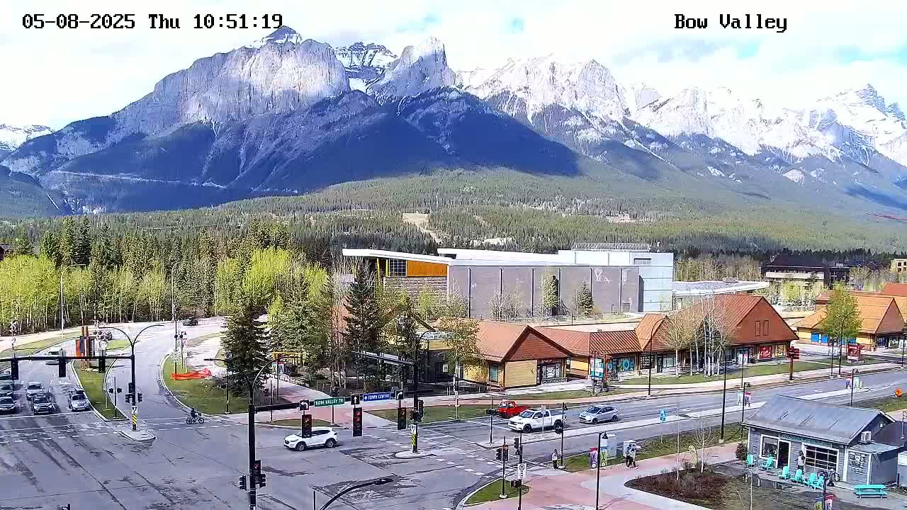 A partly sunny day reveals a mountain range capped with snow behind a town's roads and buildings, some of which are wooden and chalet-style.