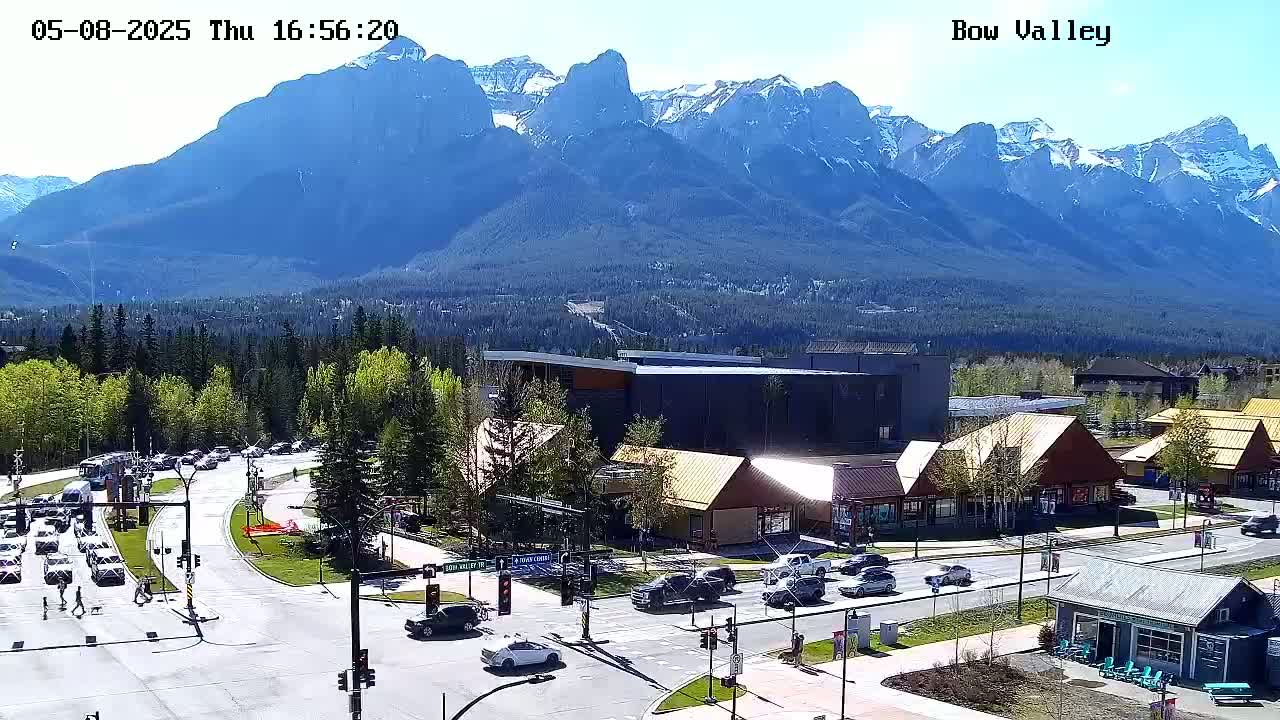 A sunny mountain vista overlooks a town square with several cars at an intersection, surrounded by low buildings and green trees.