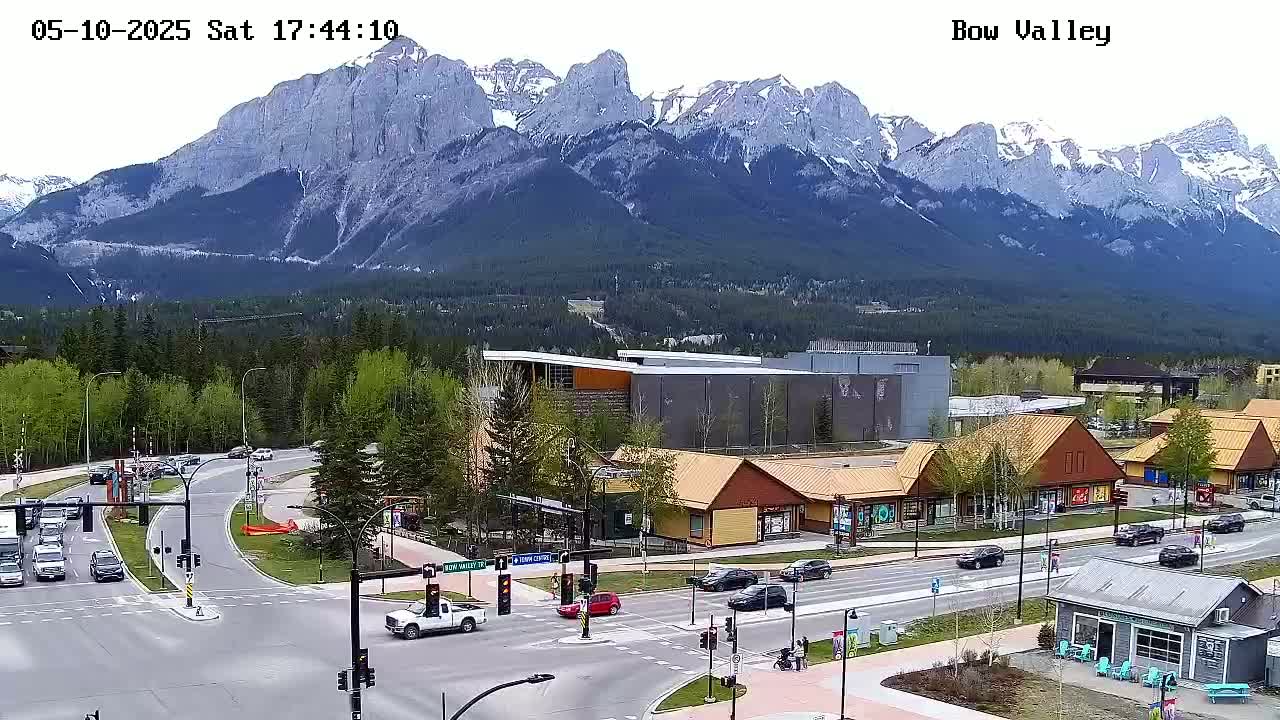 A mountain range with snow-capped peaks overlooks a town with shops and a road intersection, under a mostly clear sky.