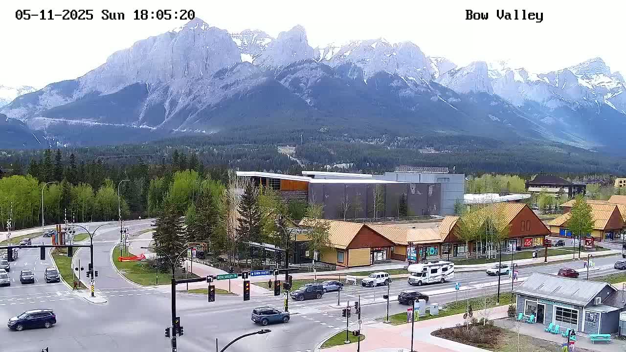 A mountain range is visible in the background of a sunny day, overlooking a town with cars driving on roads and buildings near a crossroads.