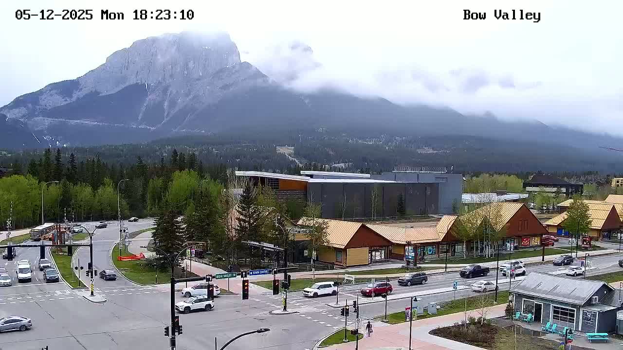 A mountain range partially obscured by clouds overlooks a town with a busy intersection and several commercial buildings.