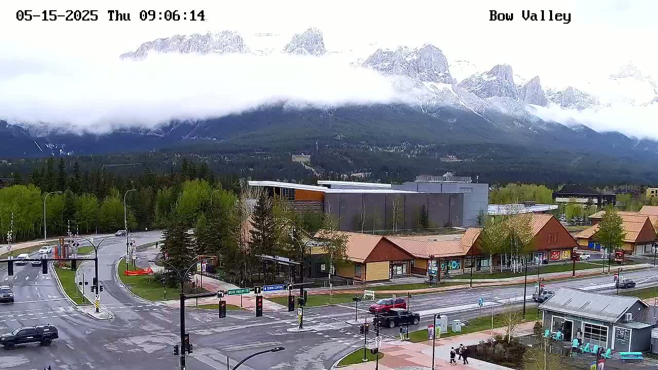 A cloudy day reveals snow-capped mountains in the distance overlooking a town intersection with several buildings and cars.
