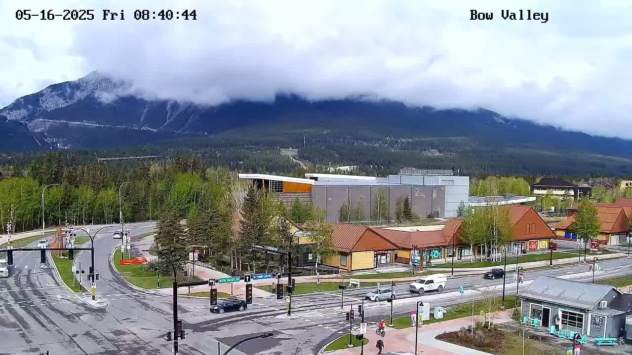 A partly cloudy day reveals a mountain range behind a town with roads, buildings, and some greenery.