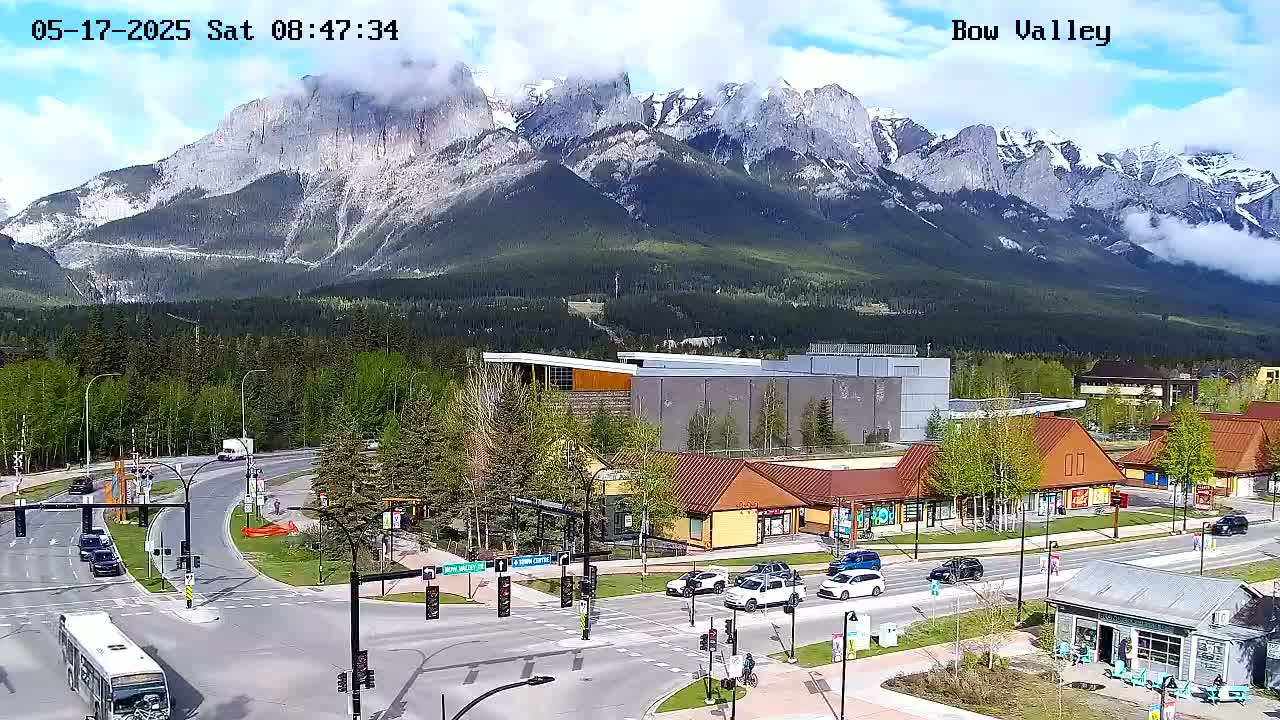 A partly cloudy day shows a mountain range in the background, overlooking a town with roads, buildings, and vehicles under a partly cloudy sky.