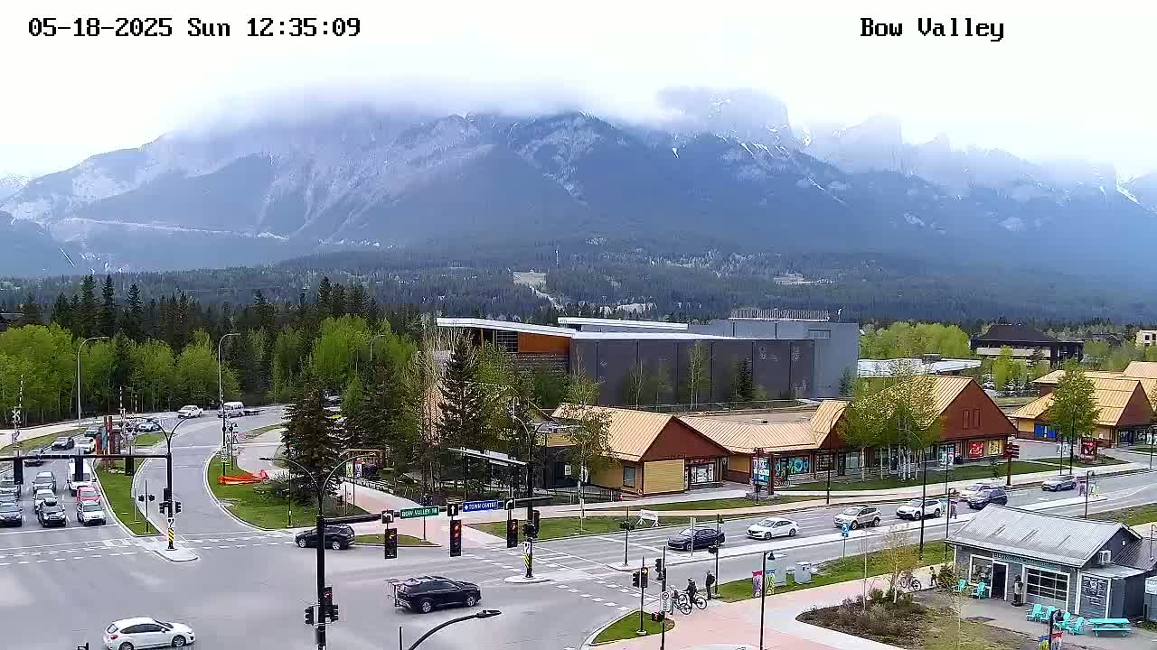 A partly cloudy day reveals a mountain range in the distance behind a town with buildings, roads, and vehicles.