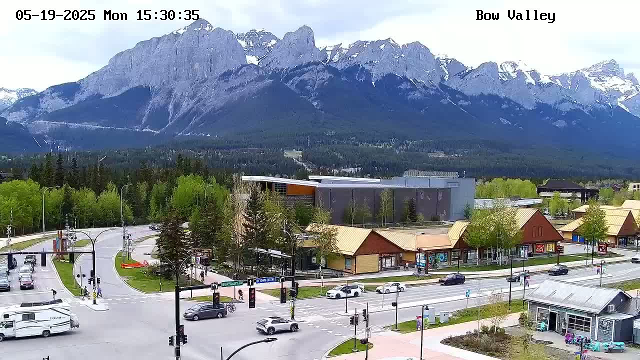 A partly cloudy day reveals a mountain range overlooking a town with a busy intersection, several shops, and green spaces.