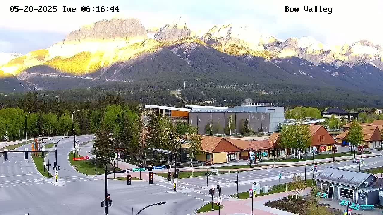 A mountain range illuminated by sunrise overlooks a town square with roads, buildings, and greenery.