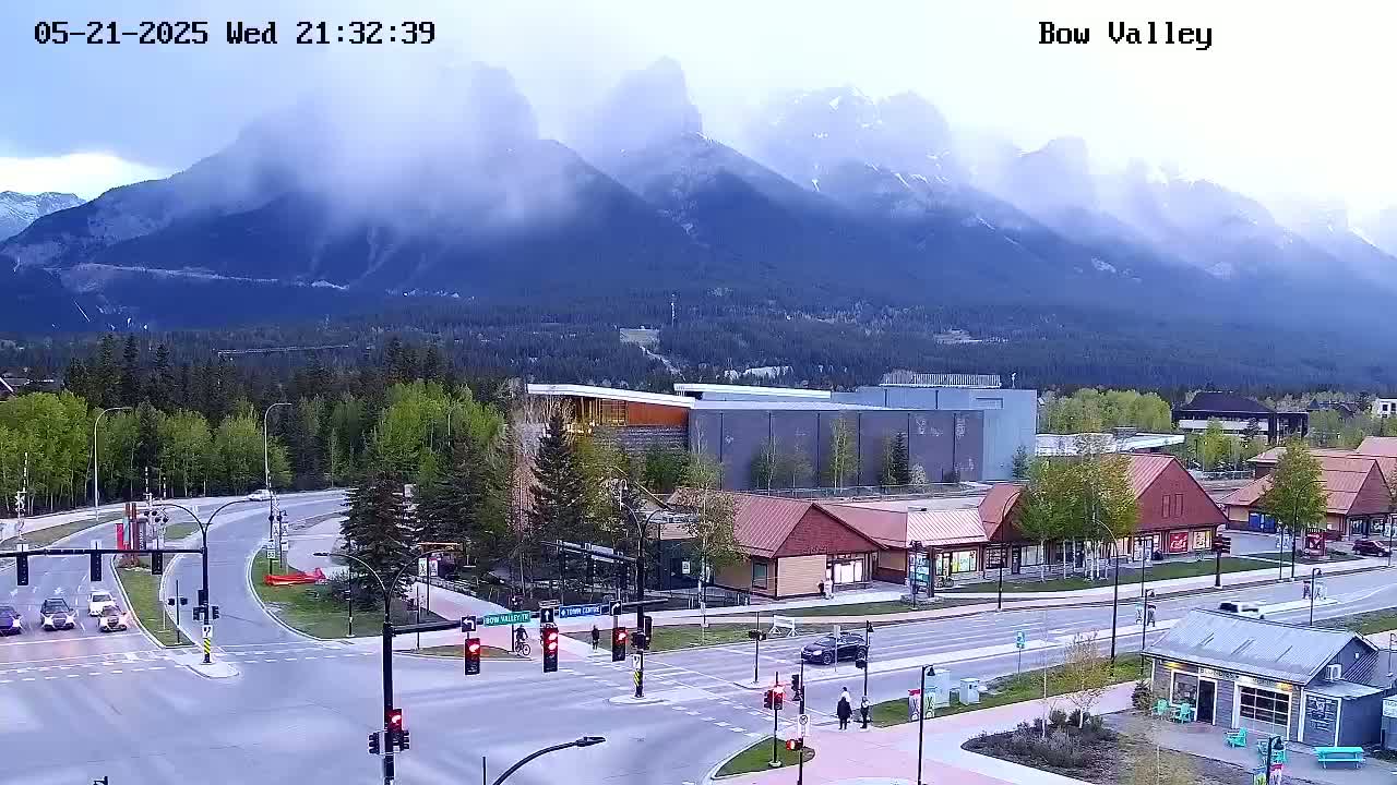 A mountain range shrouded in mist overlooks a town with roads, buildings, and sparse traffic under an overcast sky.
