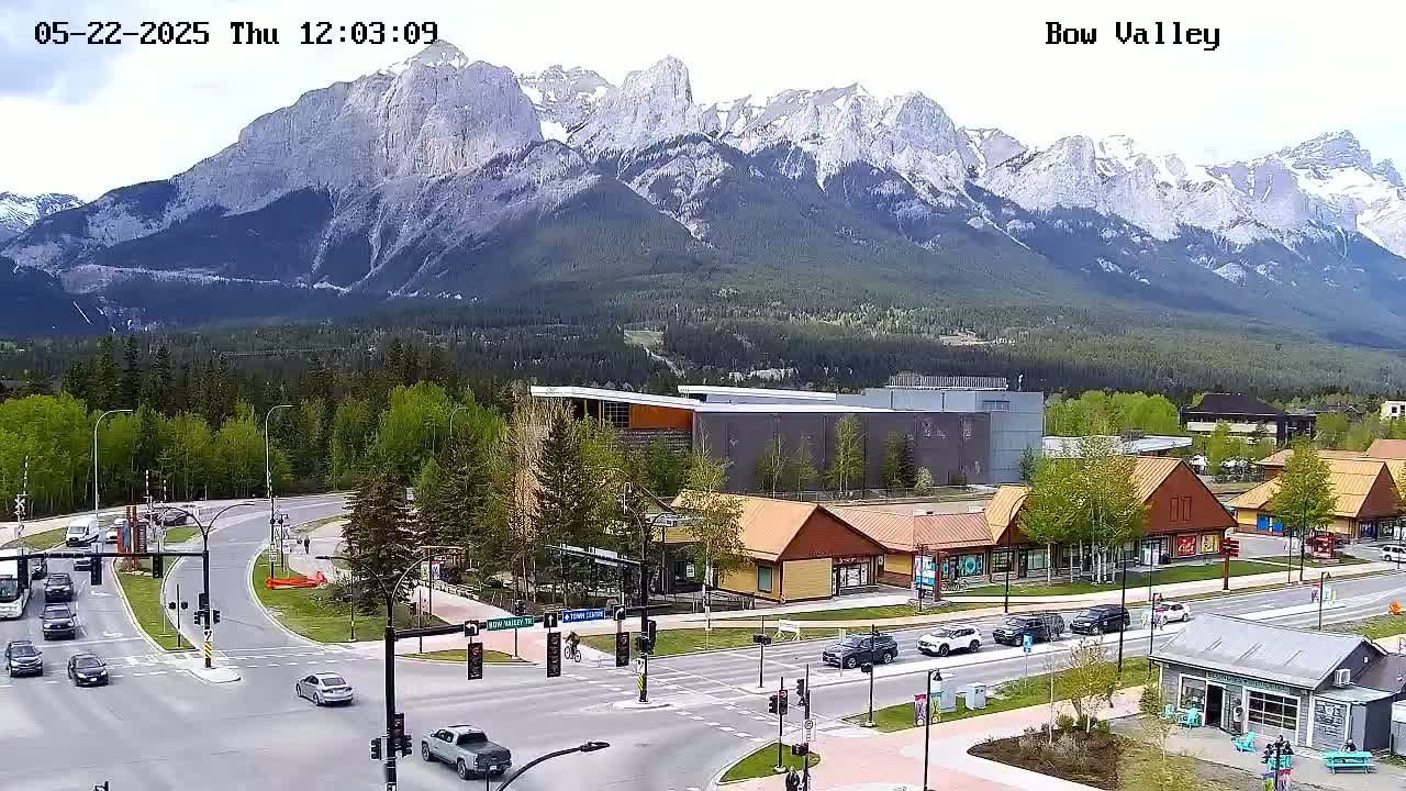A partly cloudy day reveals a mountain range in the background, overlooking a town square with buildings, roads, and vehicles.