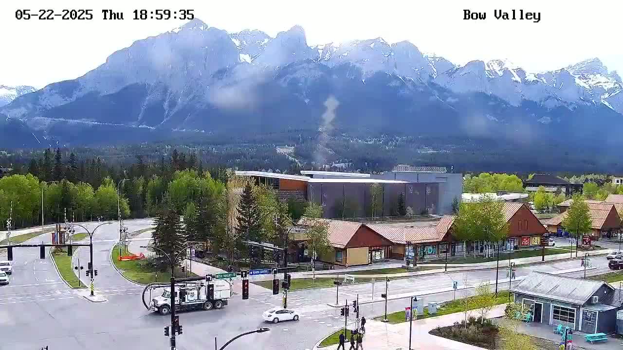 A mountain range partially covered in snow is visible in the background, with a small town and a street intersection in the foreground under partly cloudy skies.