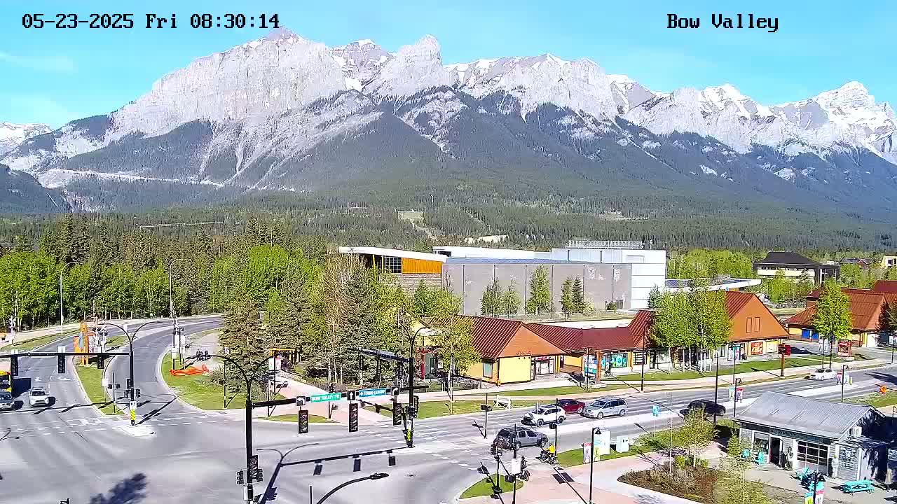 A high-angle view shows a sunny day in a mountain town with a road intersection, buildings, and snow-capped mountains in the background.