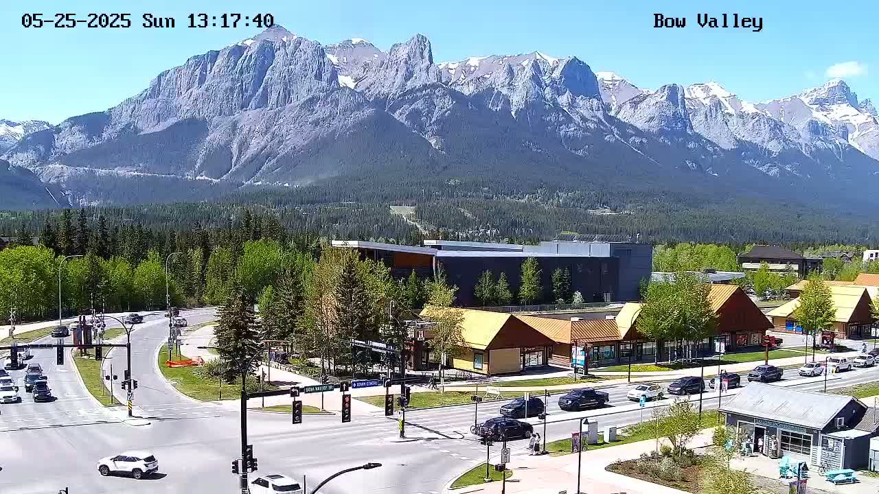 A sunny day reveals a mountain range in the background of a valley town with cars driving on roads that intersect at a traffic light, and various shops and buildings are scattered throughout the foreground.