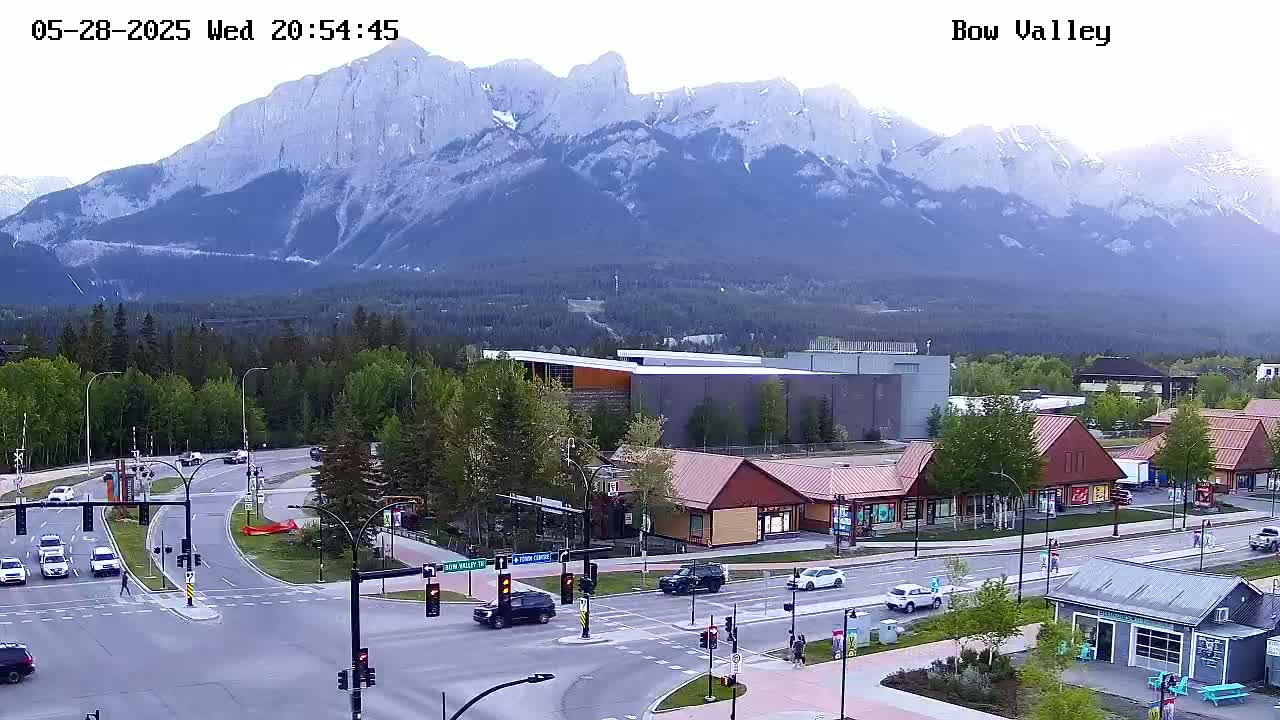A mountain range is visible in the background of a daytime scene showing a road intersection with several cars and buildings, under a mostly clear sky.