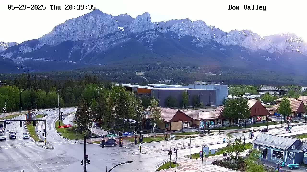 A wet road intersects with another road in front of several buildings at the base of a large mountain range under a partly cloudy sky.