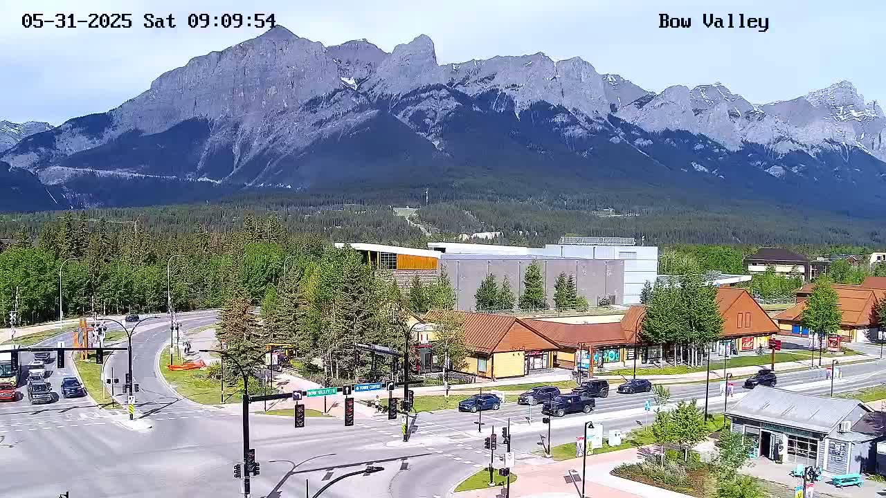 A sunny view of a mountain range behind a town with a road intersection and several buildings, including shops and a larger building.