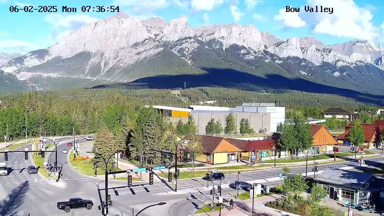 A sunny mountain vista overlooks a roadway intersection and several buildings in a town, with some cars and pedestrians visible.