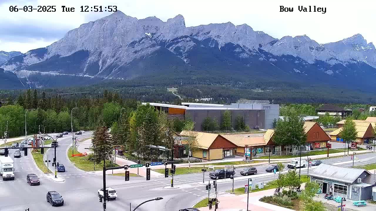 A mostly sunny day reveals a mountain range in the background, overlooking a town with a roadway intersection, several buildings, and scattered vehicles.