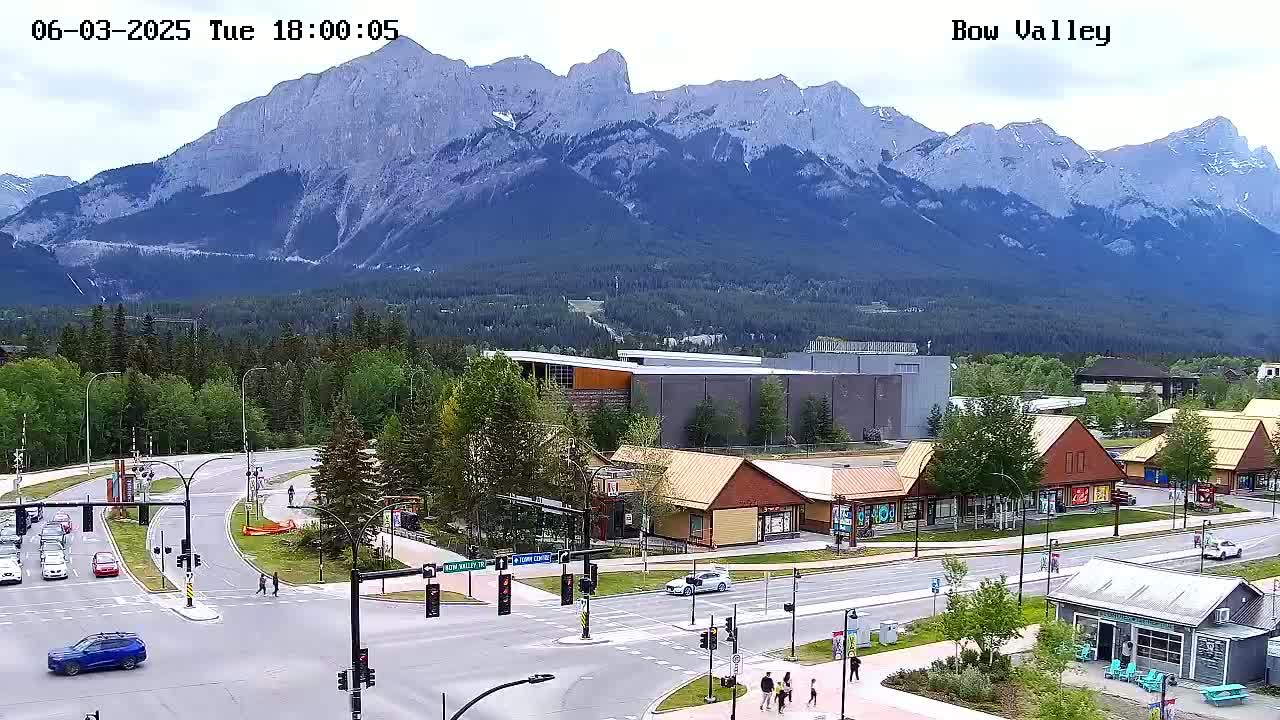 A mountain range is visible in the background of a mostly sunny outdoor scene of a town with roads, buildings, and some pedestrians.