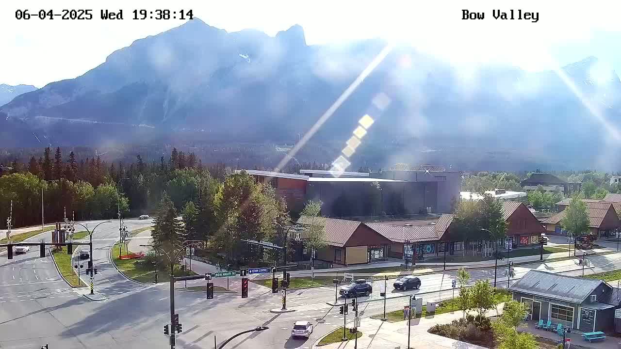 A sunny mountain view overlooks a valley intersection with several buildings and sparse traffic.