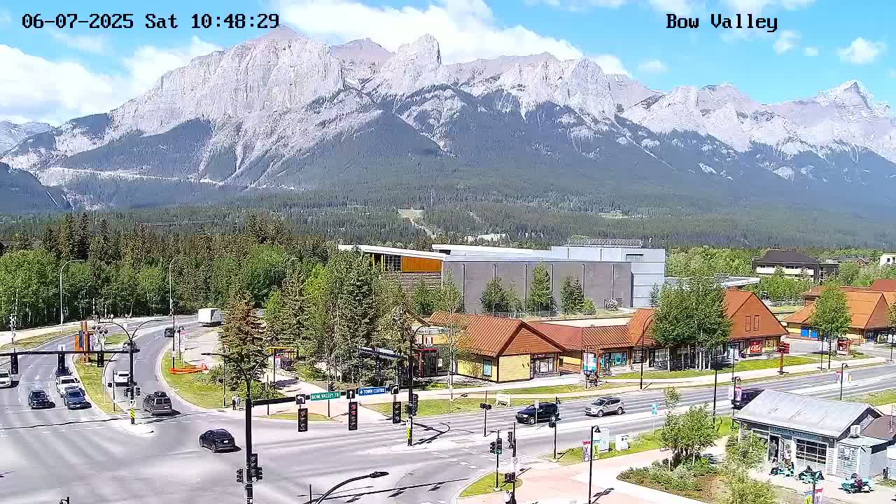 A sunny day reveals a mountain range in the background of a town with a road intersection and several buildings, including some shops and a large grey structure.