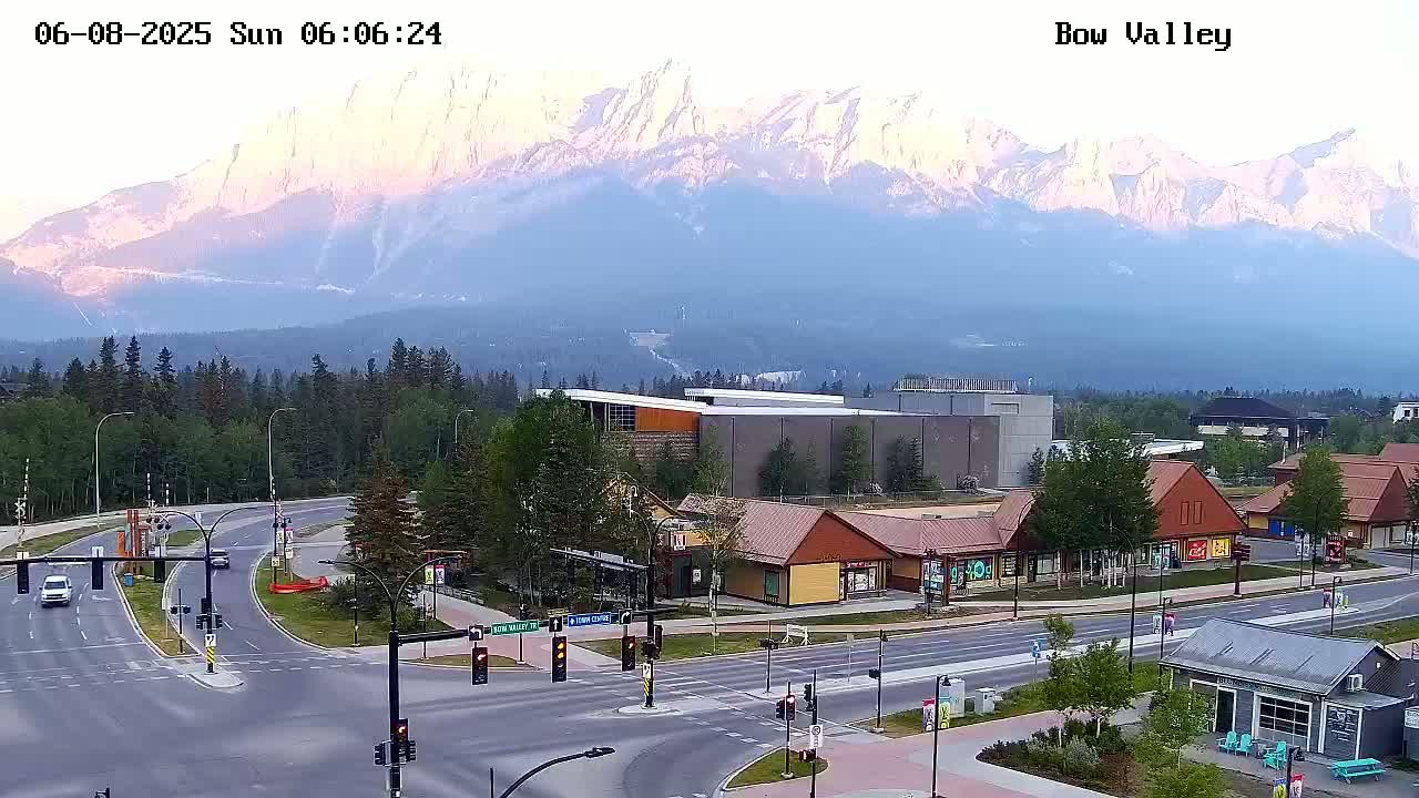 A mountain range with a pinkish hue at sunrise is visible in the distance behind a town's street intersection, buildings, and sparse traffic.