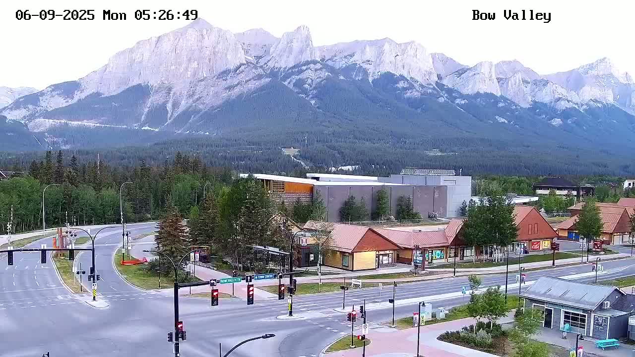 A mountain range rises behind a valley town with roads, buildings, and green spaces, under a clear sky.