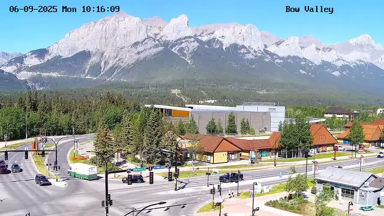 A sunny day reveals a mountain range in the background of a town with a road intersection, several buildings, and sparse traffic.