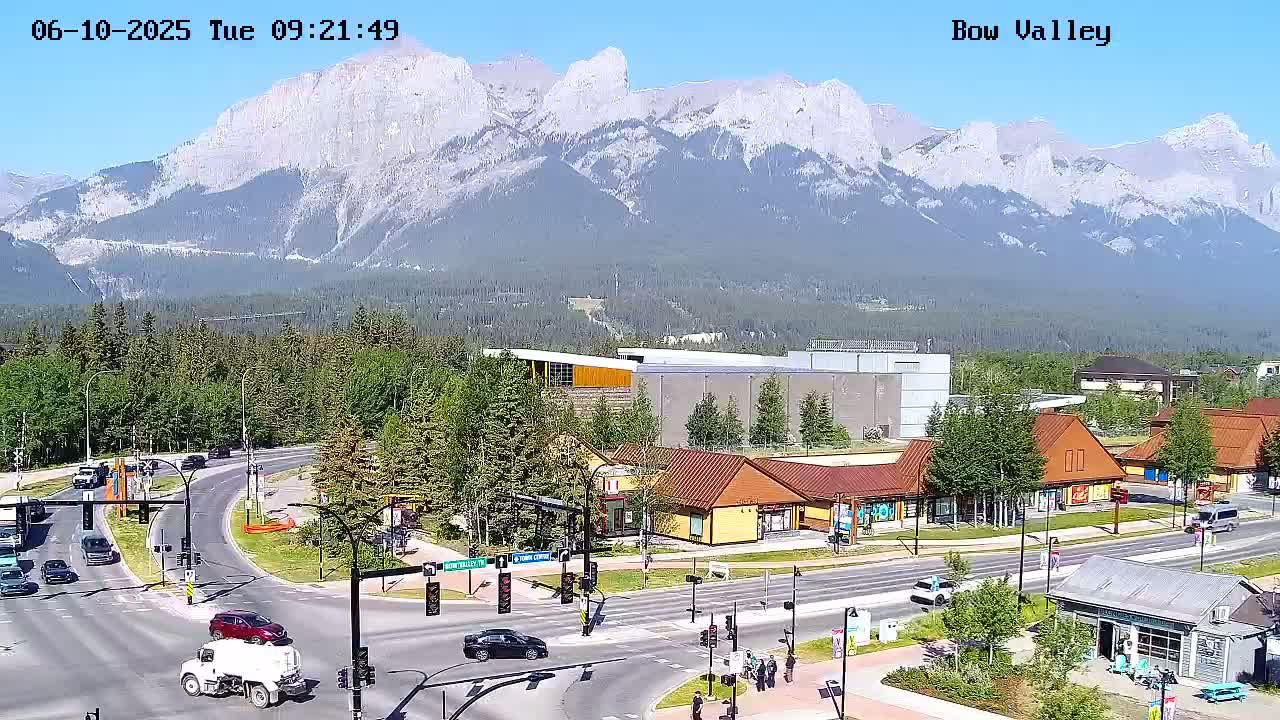 A sunny day reveals a mountain range in the background of a town's intersection with buildings and several cars.