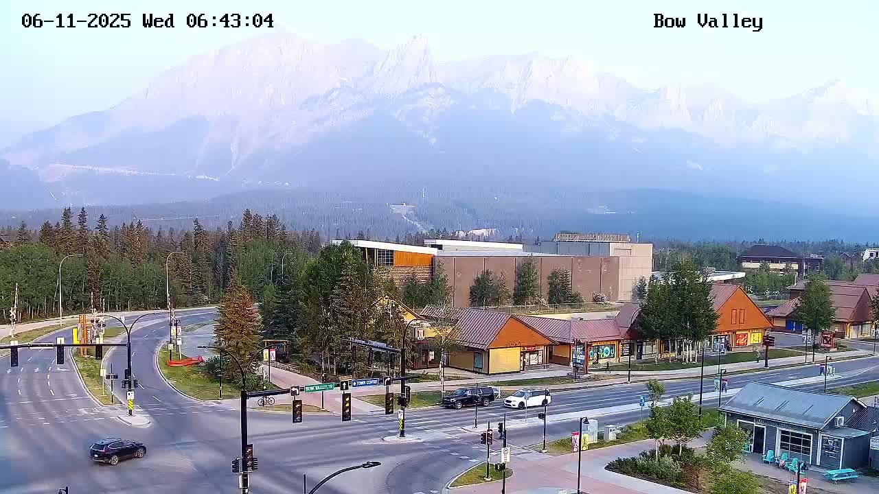 A hazy, mountain-backed town scene shows a multi-lane road intersection with several cars and buildings, including small shops and a larger building, all under a mostly clear sky.