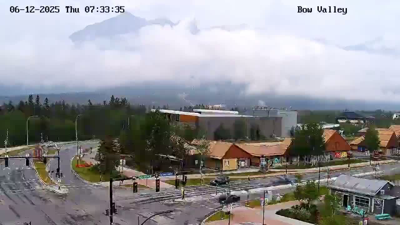 A cloudy day reveals a mountain range in the distance, overlooking a road intersection and small buildings in a town.