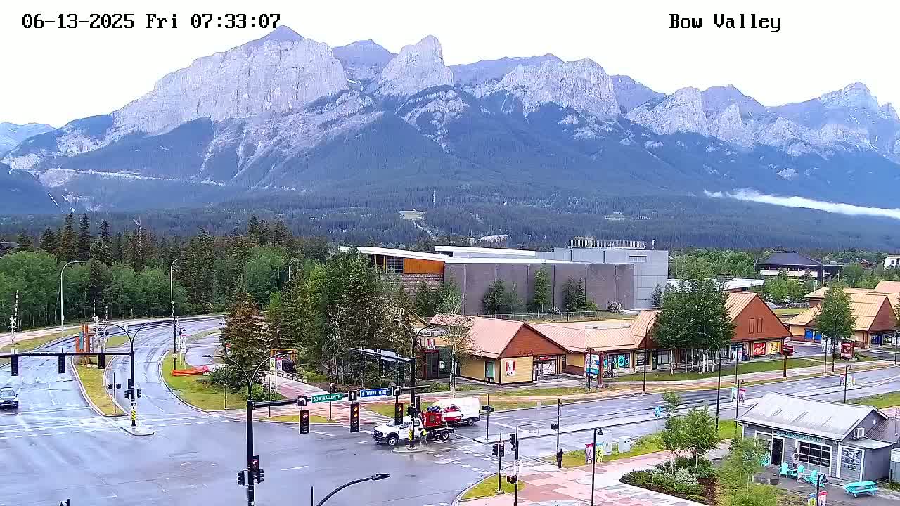A wet street intersection with surrounding buildings and a mountain range in the background under overcast skies.