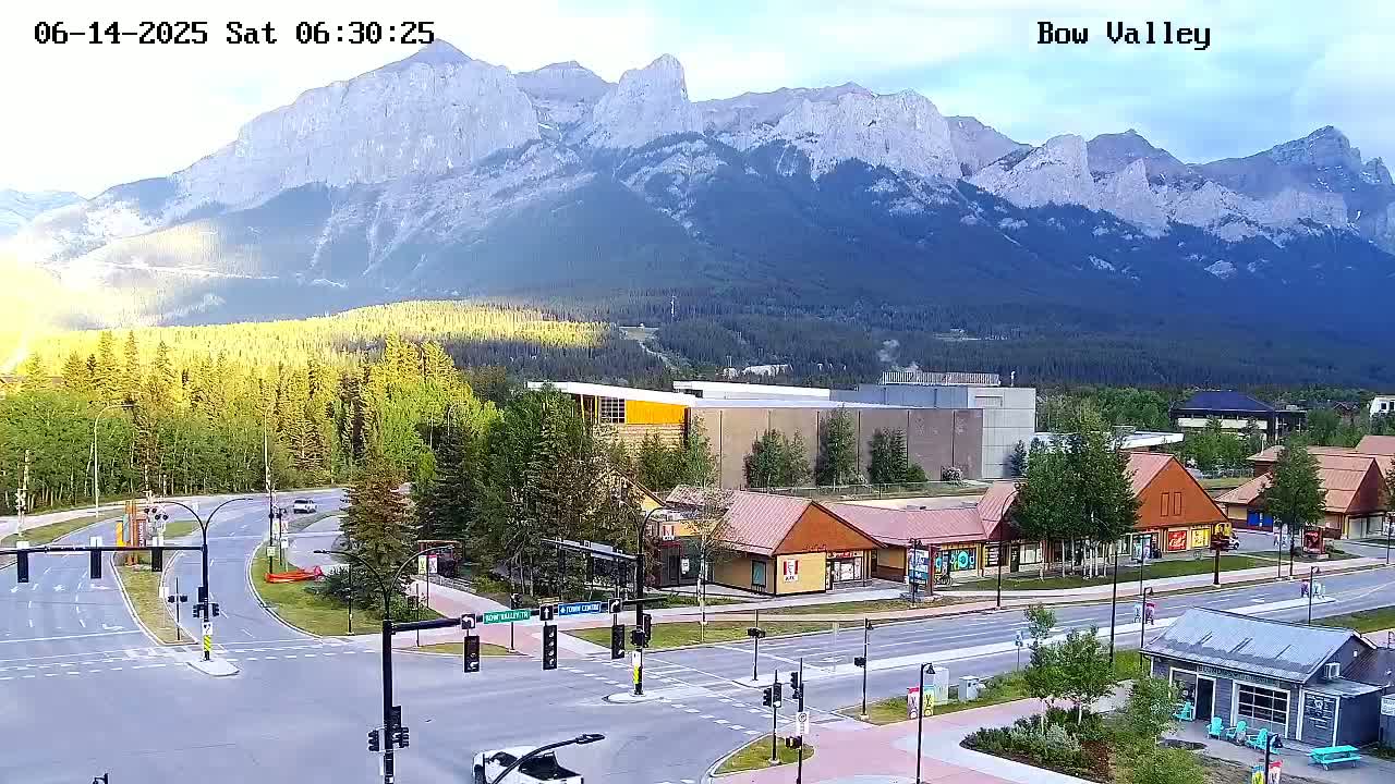 A high-angle, long shot of a mountain town at sunrise, featuring a paved intersection with several buildings and a large mountain range in the background.