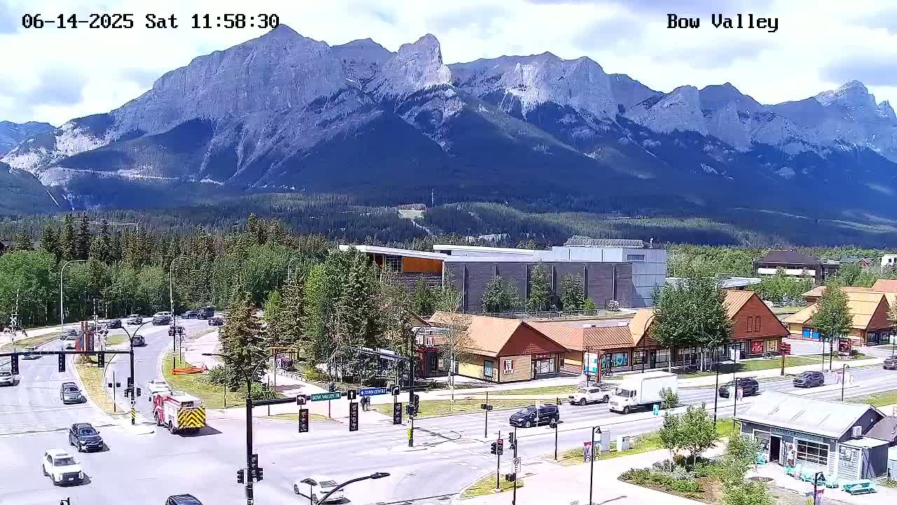 A partly cloudy day shows a mountain range in the background overlooking a town with traffic at an intersection and several buildings of various styles.