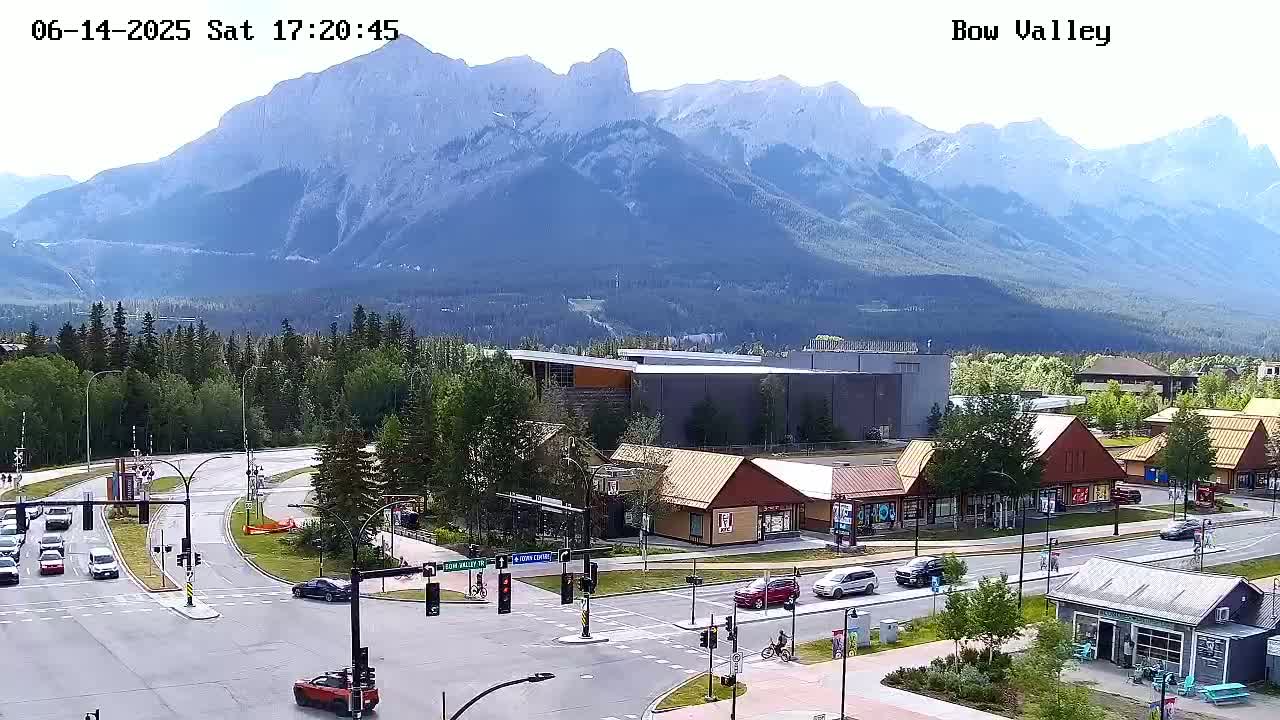A partly cloudy day shows a mountain range in the background overlooking a town with traffic at an intersection and several buildings of various styles.