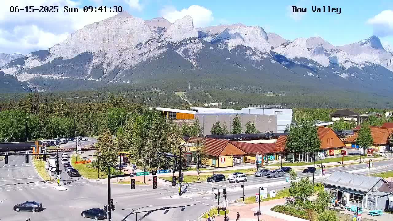 A sunny day in a mountain town shows a busy intersection with cars and pedestrians, in front of various buildings and shops, with a large mountain range in the background.