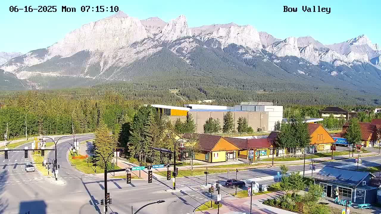 A high-angle view of a mountain town intersection with several buildings and a mountain range in the background on a clear, sunny day.