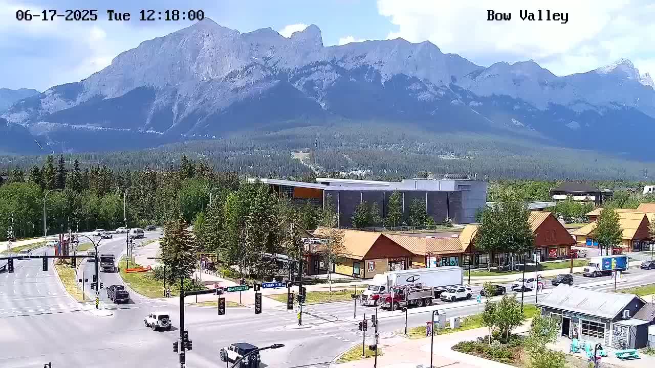 A sunny day shows a mountain range in the background, with a town featuring a road intersection and various buildings, including shops and what appears to be a recreation center.