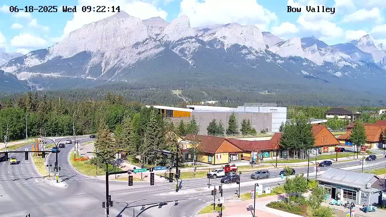 A partly cloudy day reveals a mountain range behind a town with a road intersection and several small buildings, some cars are visible on the road.