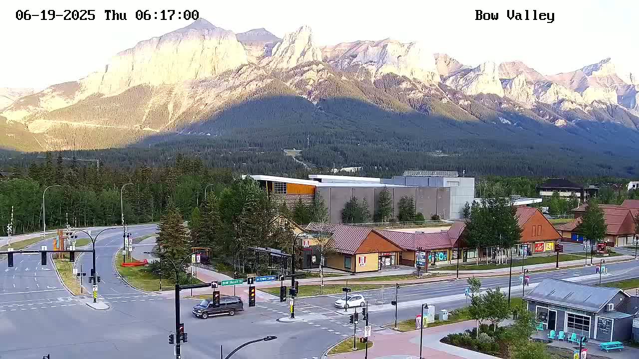 A mountain range bathed in the golden light of sunrise overlooks a town intersection with several buildings and vehicles.