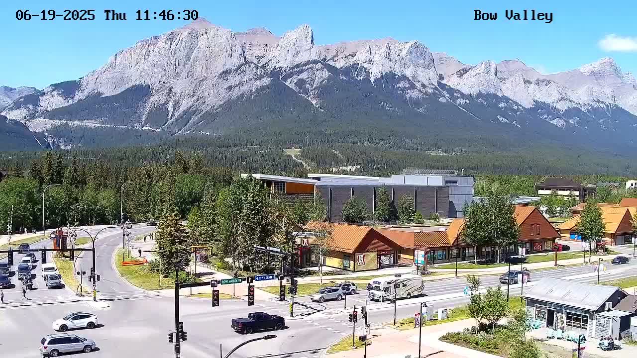 A sunny mountain view overlooks a town with several buildings and cars at a crossroads.
